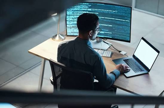 A developer working from behind at a desk with a wide curved monitor full of code and a laptop